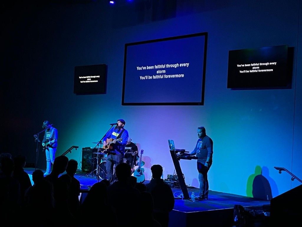 Band performing under blue stage lights with lyrics on screens.