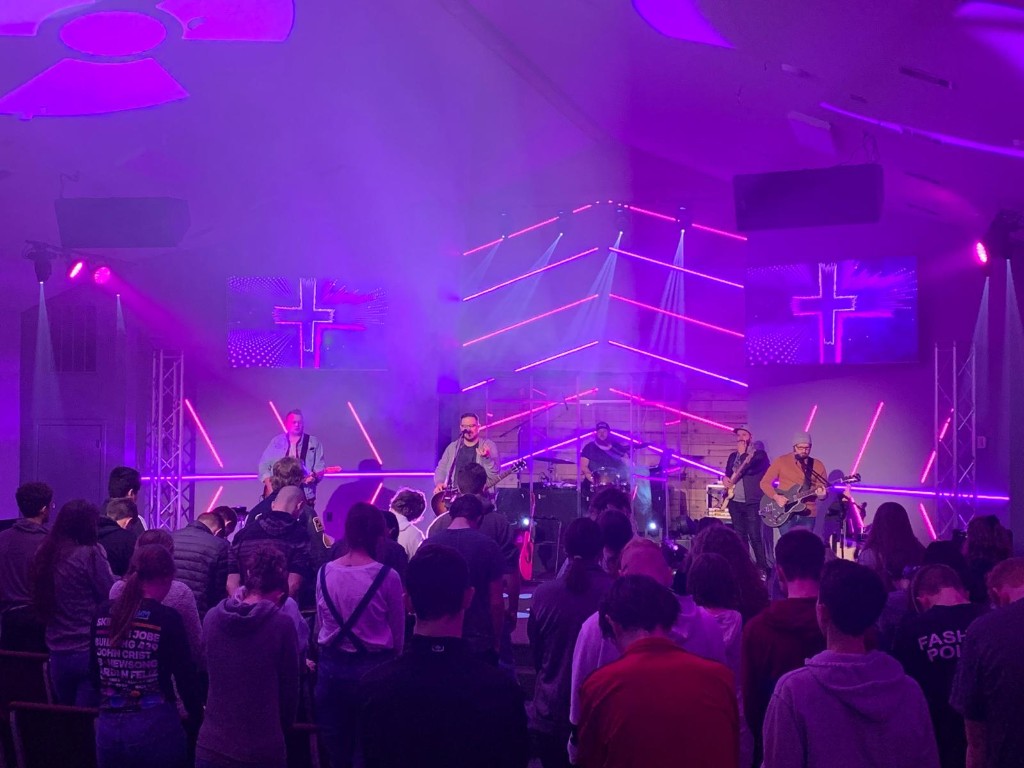Live band on stage with purple lighting and crosses on LED screens.
